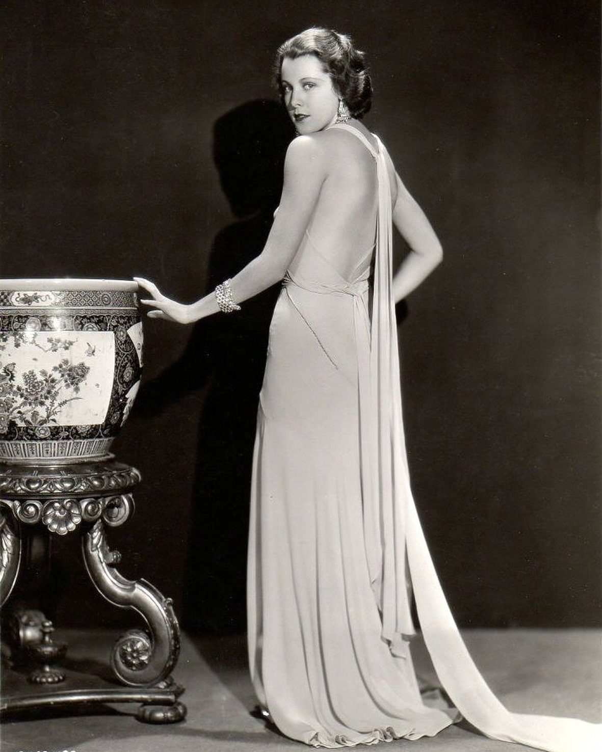 Woman in a backless vintage evening gown posing next to an ornate vase.