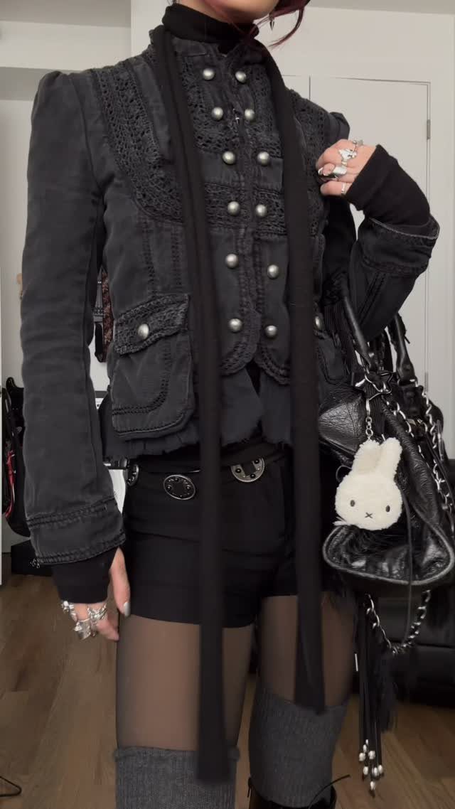 Person wearing a cropped black gothic jacket with silver buttons and embroidery.