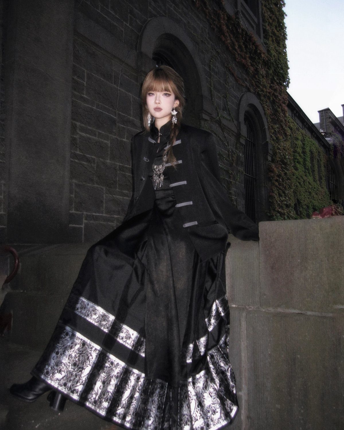Person in layered black gothic outfit posing by a stone building with ivy.