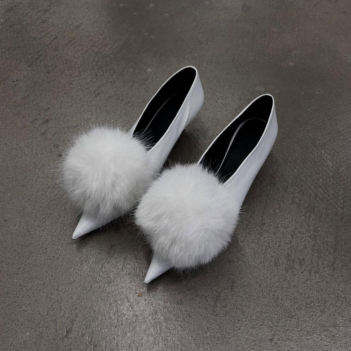 White pointed-toe pumps with large white fluffy pom-pom charms on the toes.