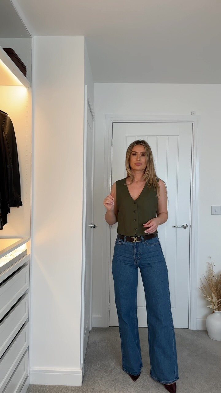 Woman wearing olive green sleeveless vest and high-waisted wide-leg blue jeans in a room.