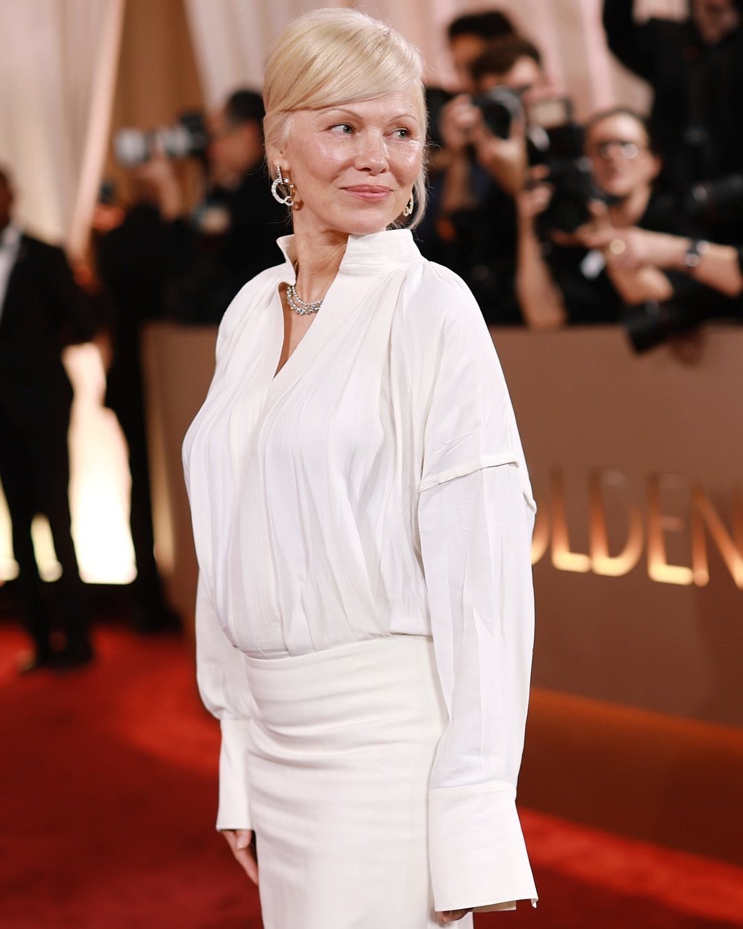 Pamela Anderson in a white blouse and skirt on a red carpet.