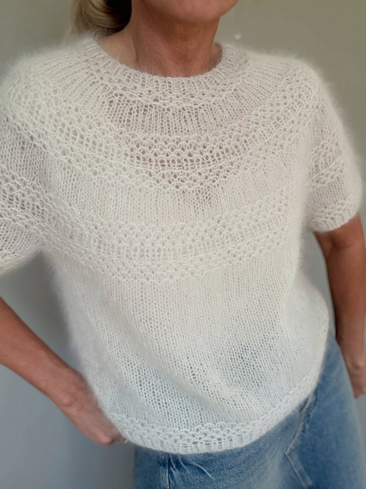 Woman wearing a white short-sleeved knitted top with lace detailing and blue jeans.