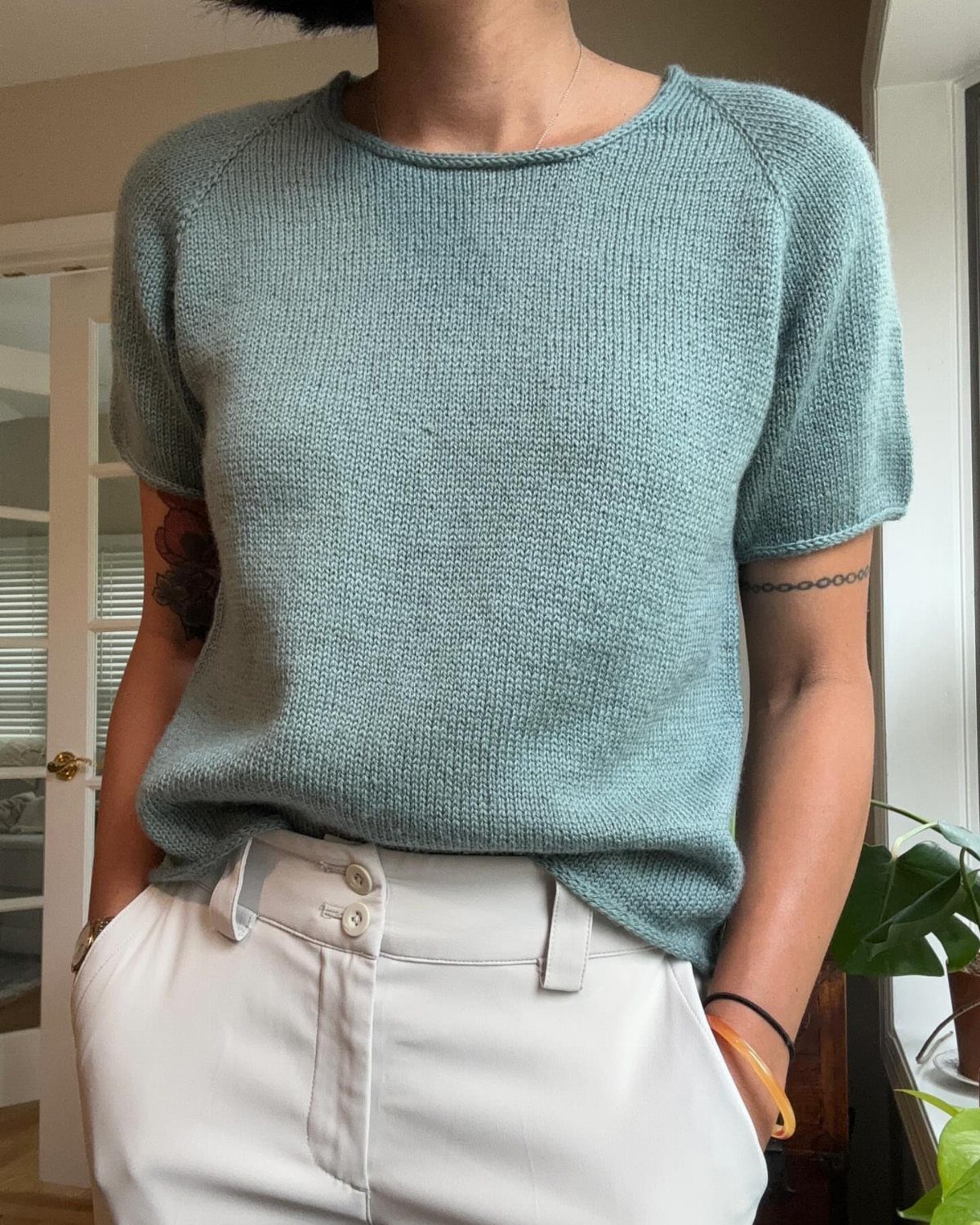 Light teal short-sleeved knitted top paired with high-waisted white trousers indoors.