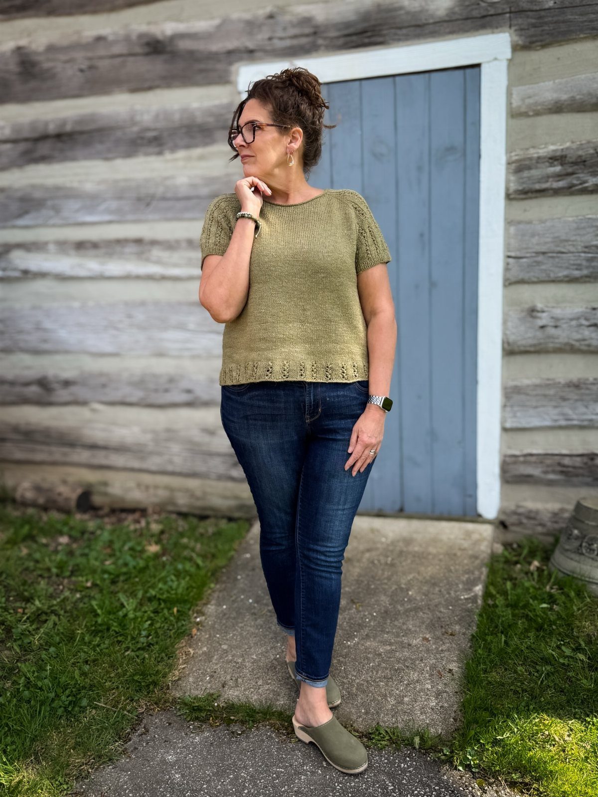 Woman wearing an olive-green knitted short-sleeved top and dark blue jeans outdoors.