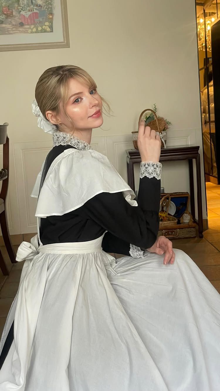Woman in a black long-sleeved dress with a white apron and lace trim.