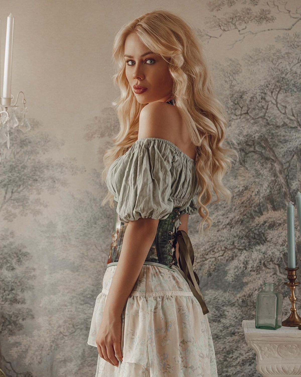Blonde woman in a medieval blouse, corset, and long white skirt posing indoors.