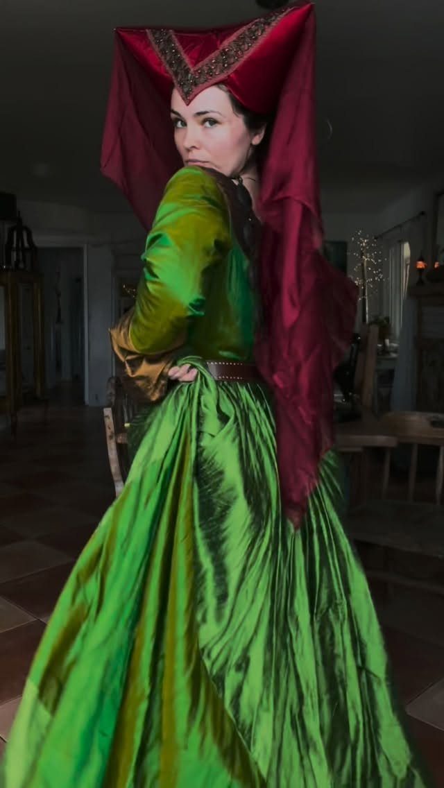 Woman in a floor-length green medieval dress and a tall red hennin headpiece.