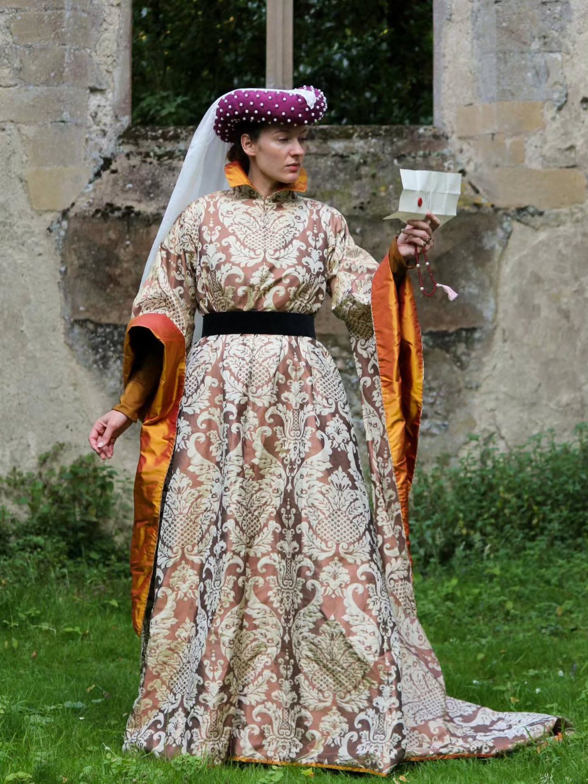 Woman in a gold patterned medieval noble dress and purple jeweled headdress outdoors.