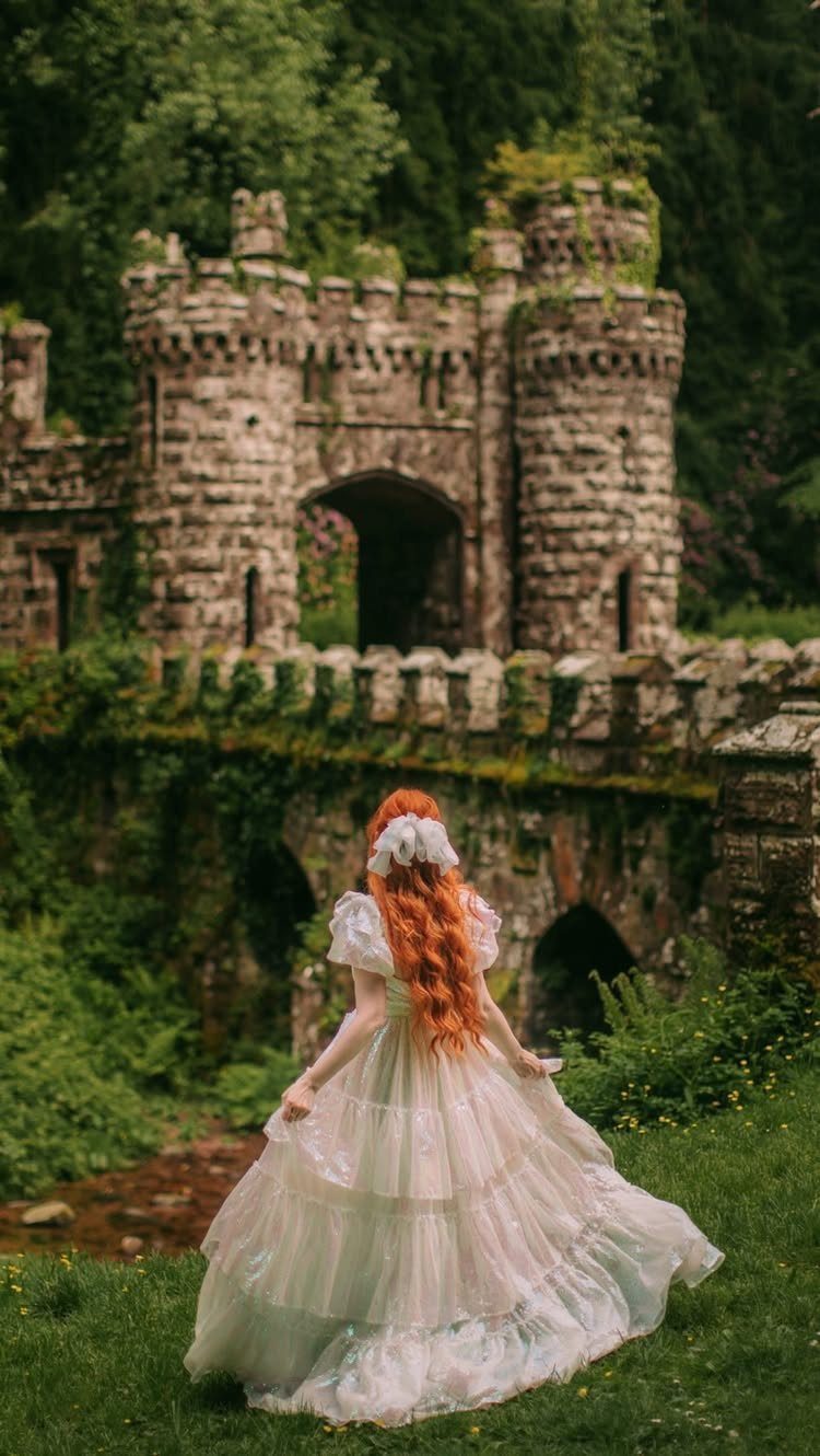 Woman in a white tiered medieval gown standing before a stone castle ruin.