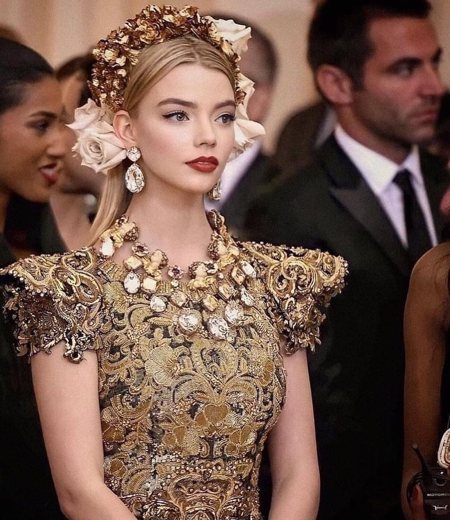 Woman in an ornate gold embroidered gown and floral headpiece at a gala.