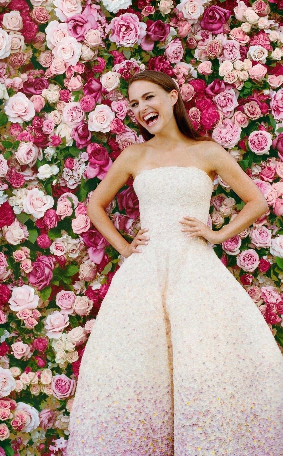 Woman in strapless white floral gown laughing before a wall of pink roses