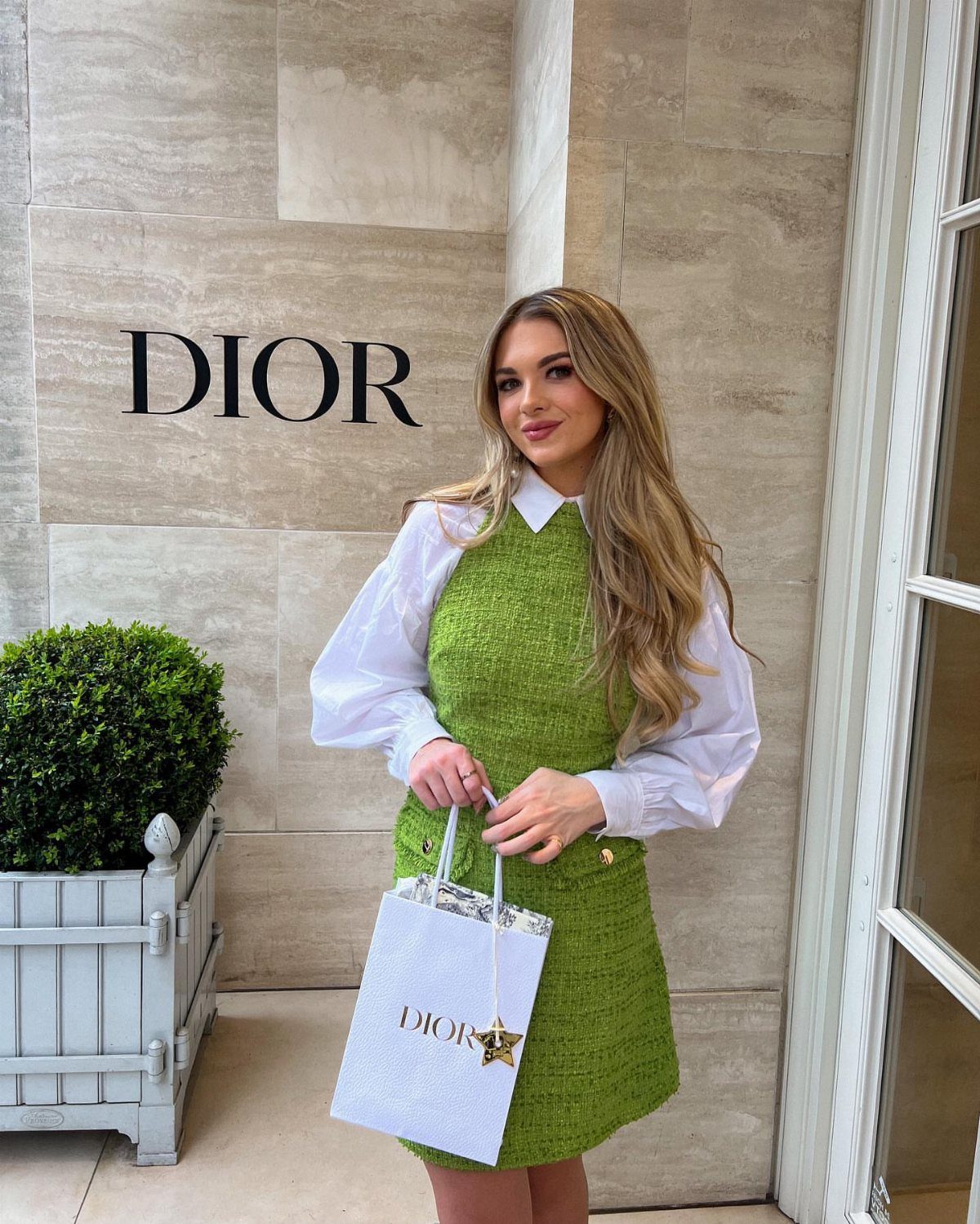 Woman in green tweed mini dress and white shirt holding Dior shopping bag.