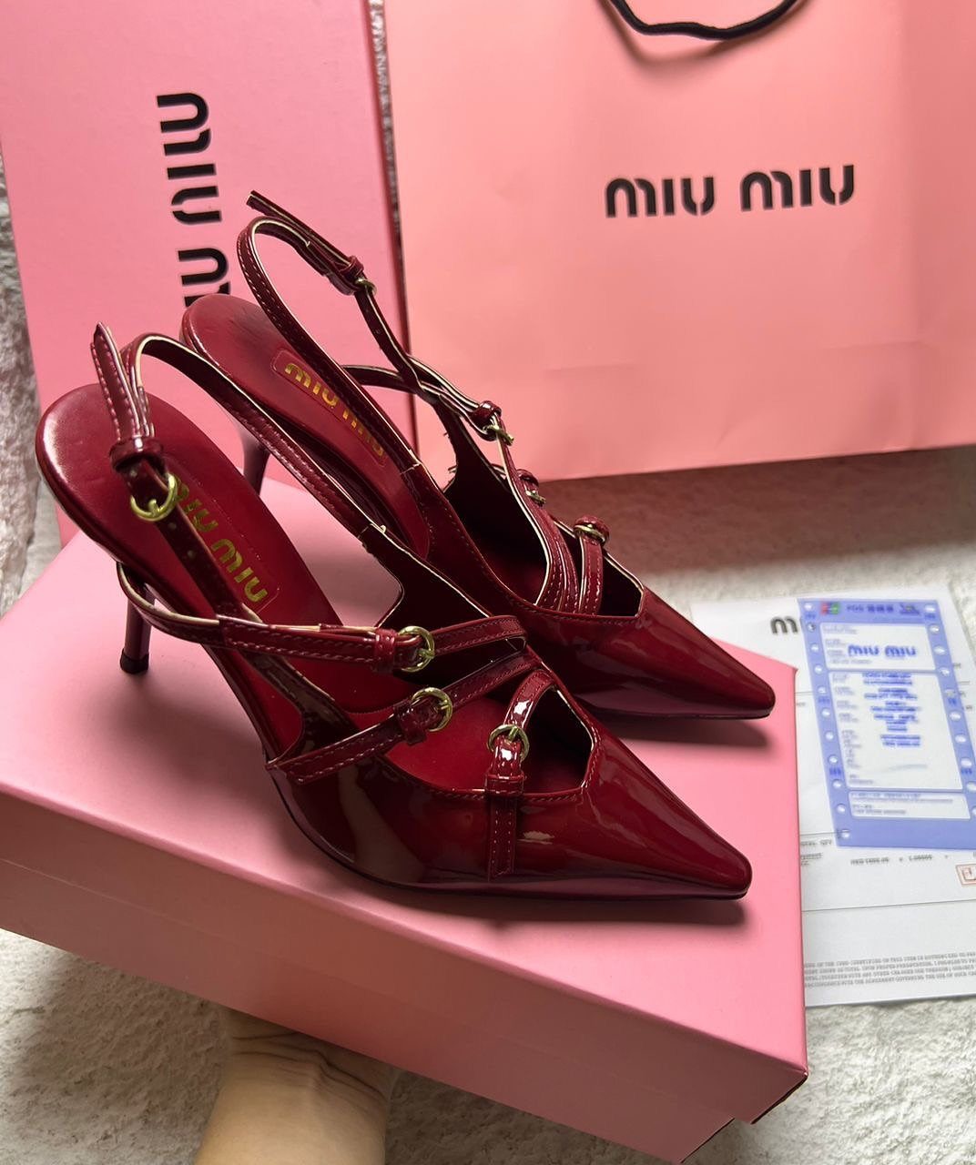 Burgundy glossy Miu Miu pointed-toe heels on a pink shoebox with shopping bag.