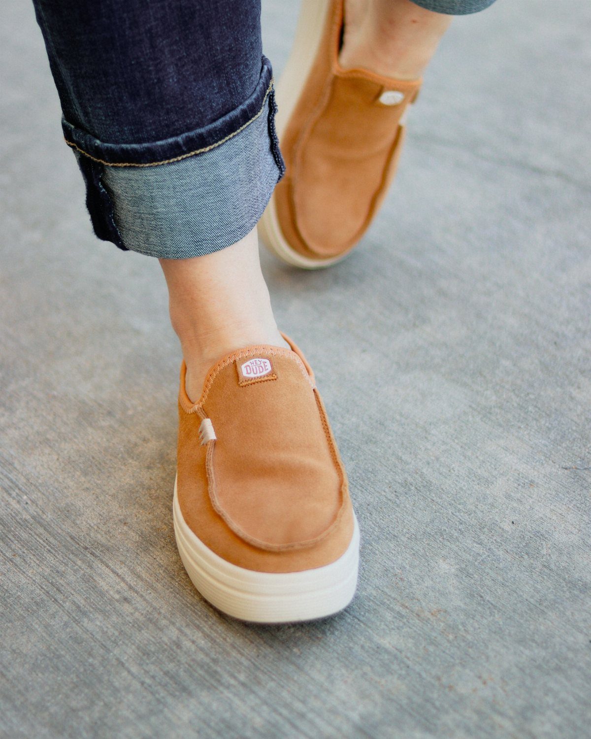 Tan suede slip-on shoes with white soles worn with cuffed blue jeans.