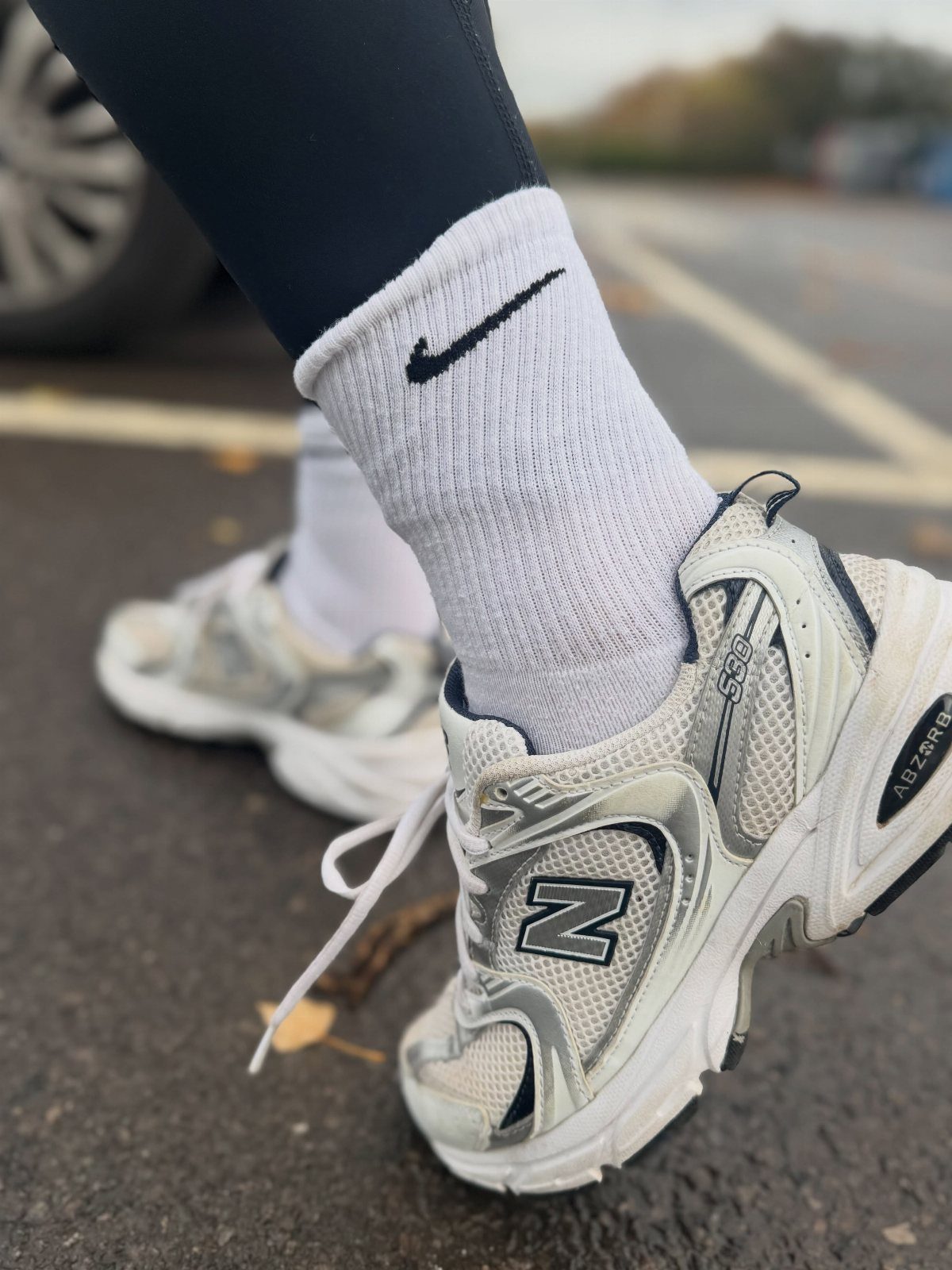 White and silver New Balance 530 sneakers with white Nike socks and black leggings.
