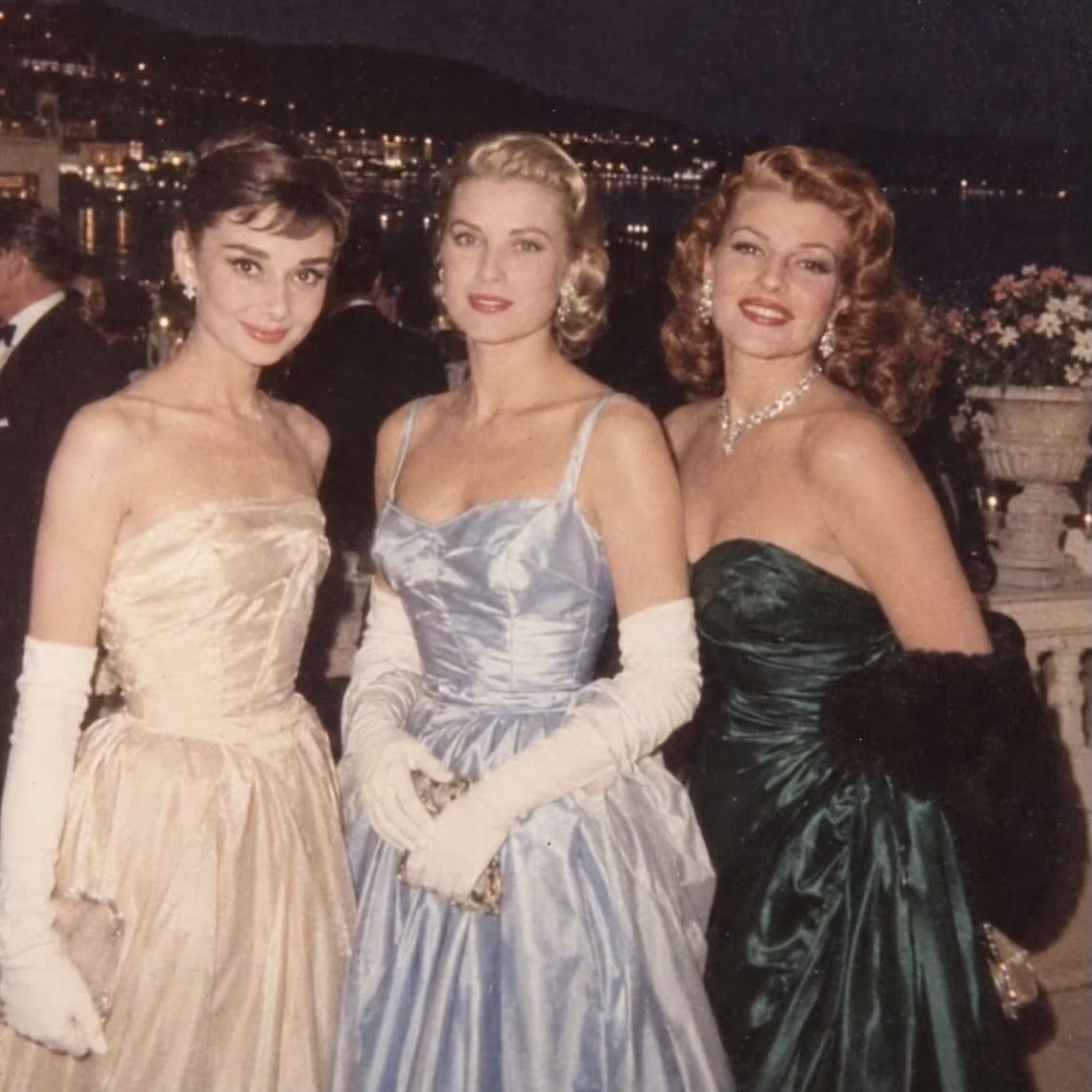 Three women in satin vintage evening gowns and white gloves posing at night.