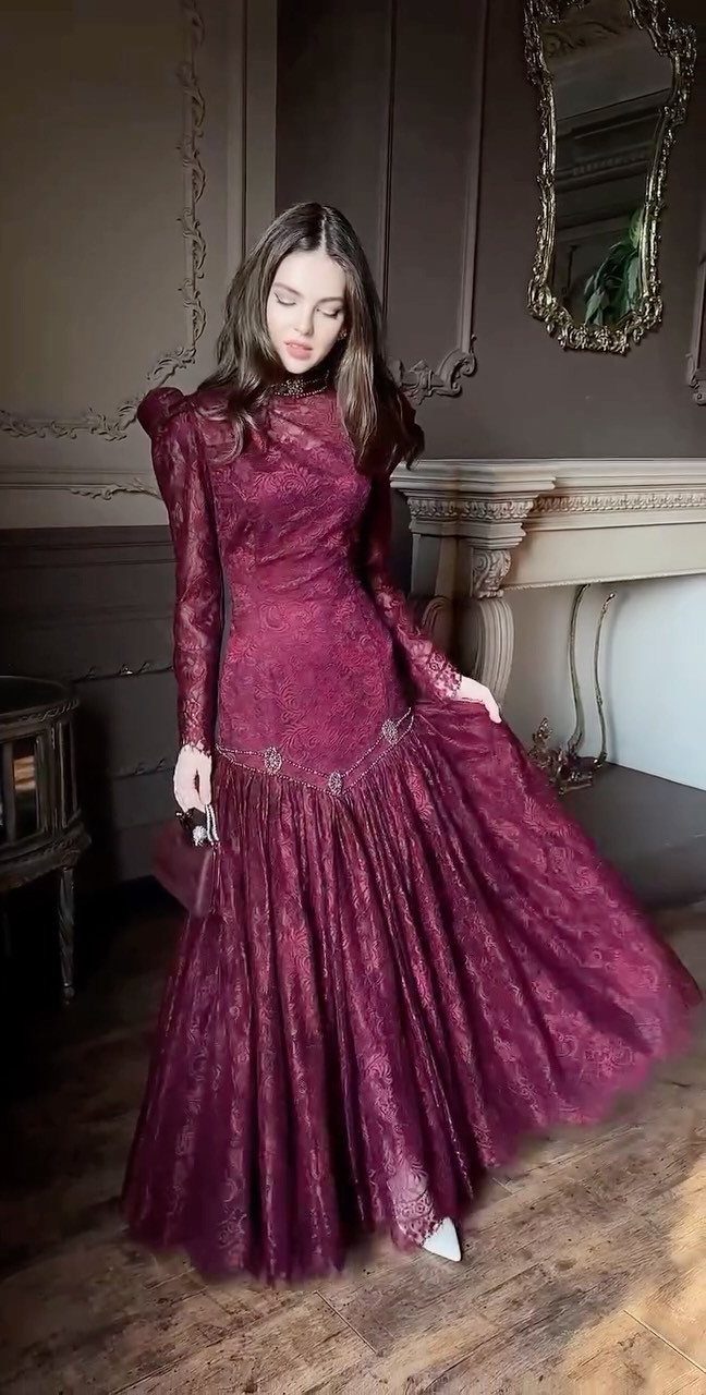 Woman in a long-sleeved burgundy lace gown standing in a vintage room.