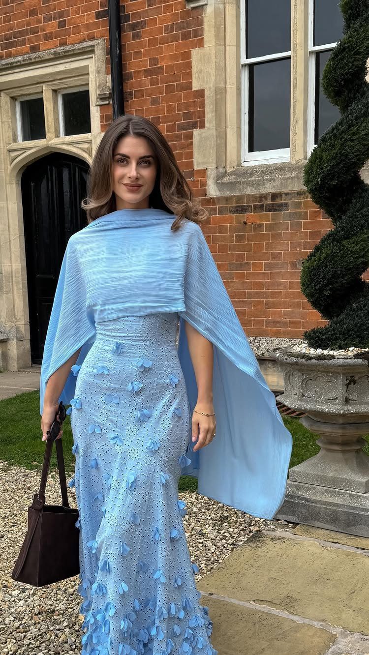 Woman in a light blue floral textured gown and capelet standing by brick building