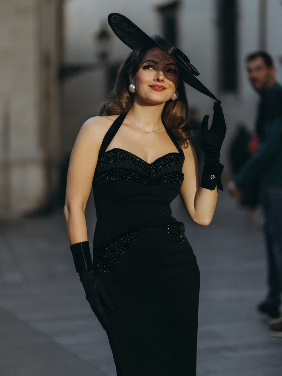 Woman in a black halter-neck dress, black hat, and gloves posing outdoors.