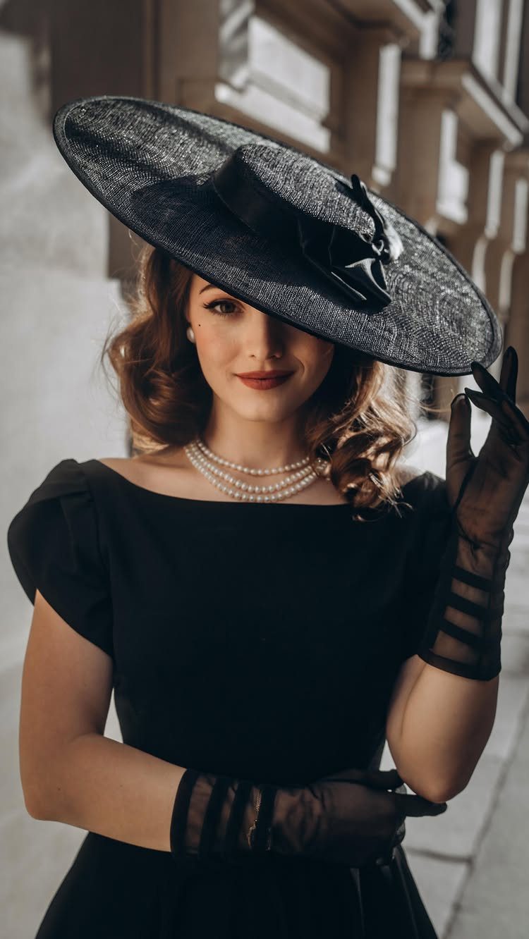 Woman in a black vintage dress, wide-brimmed hat, and pearls posing outdoors.