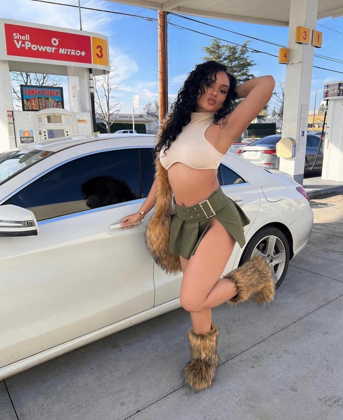Woman in beige crop top and short olive green skirt posing by white car