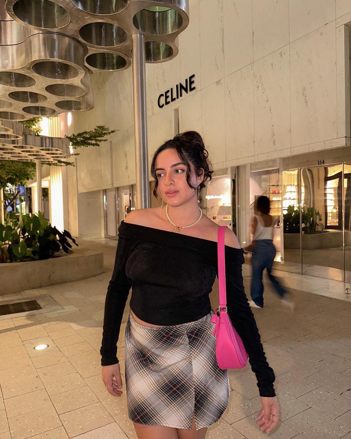 Woman in black off-the-shoulder top and plaid mini skirt with pink shoulder bag.