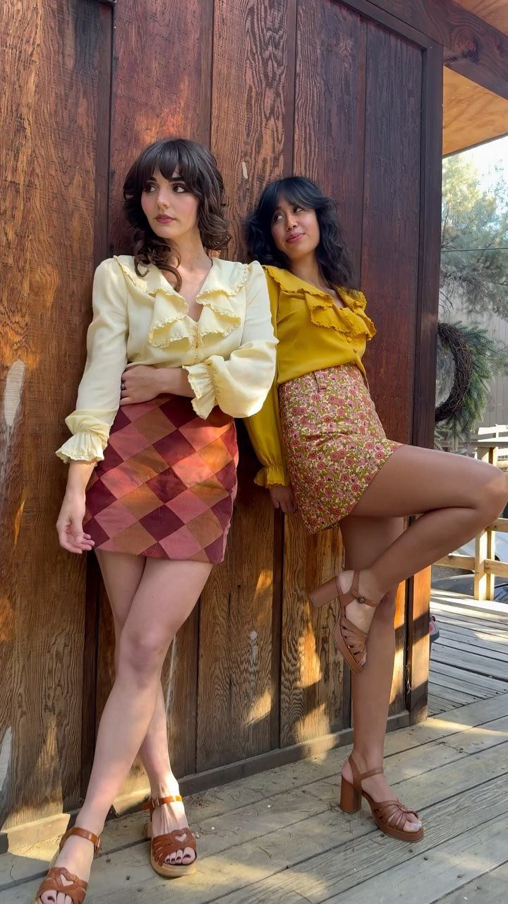 Two women wearing ruffle blouses and short skirts posing against a wooden wall.