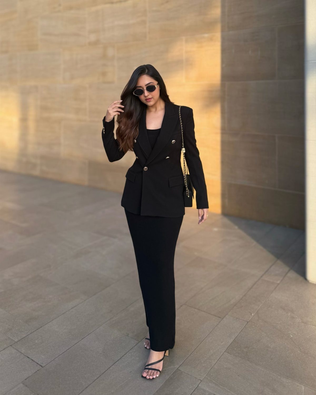 Woman in a black blazer and long black dress standing by a stone wall.