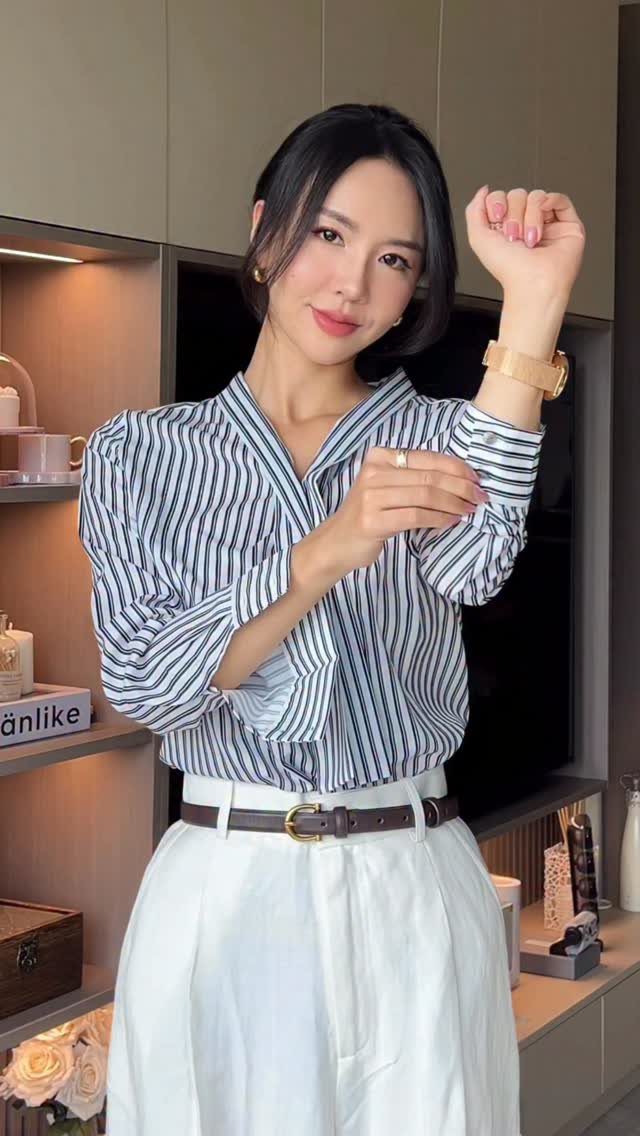 Woman in a striped button-down shirt and white trousers posing in a room.