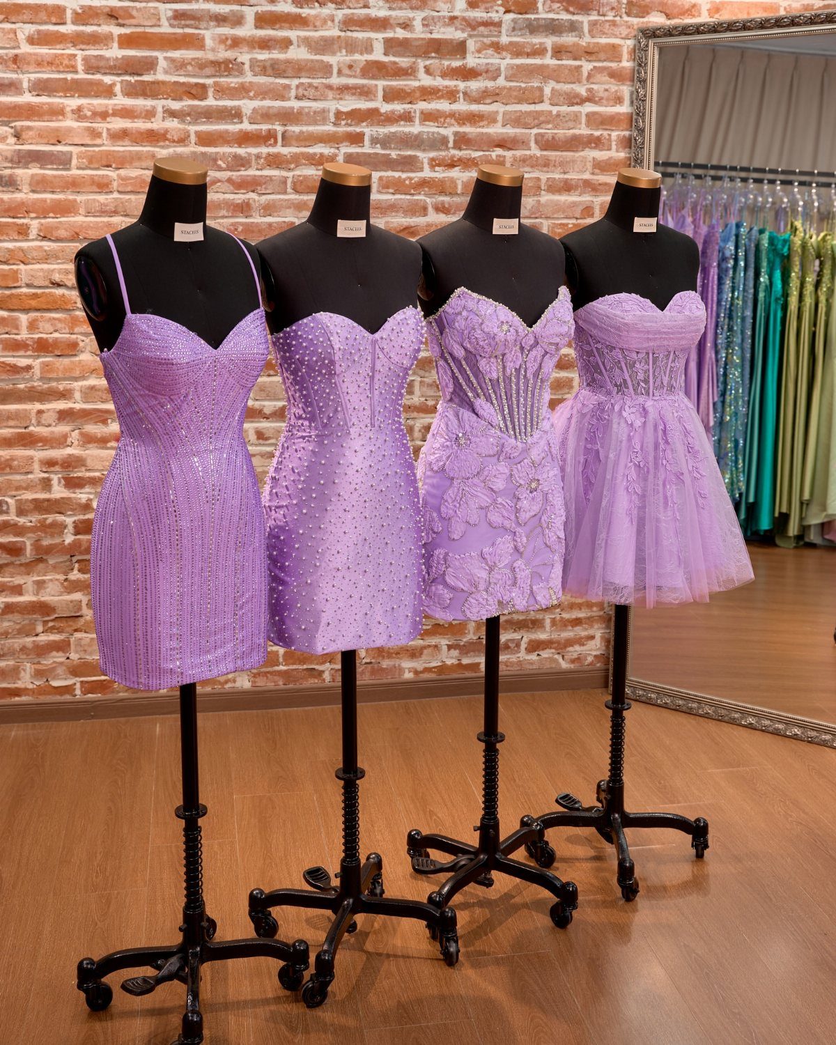 Four purple homecoming dresses on mannequins displayed against a red brick wall.