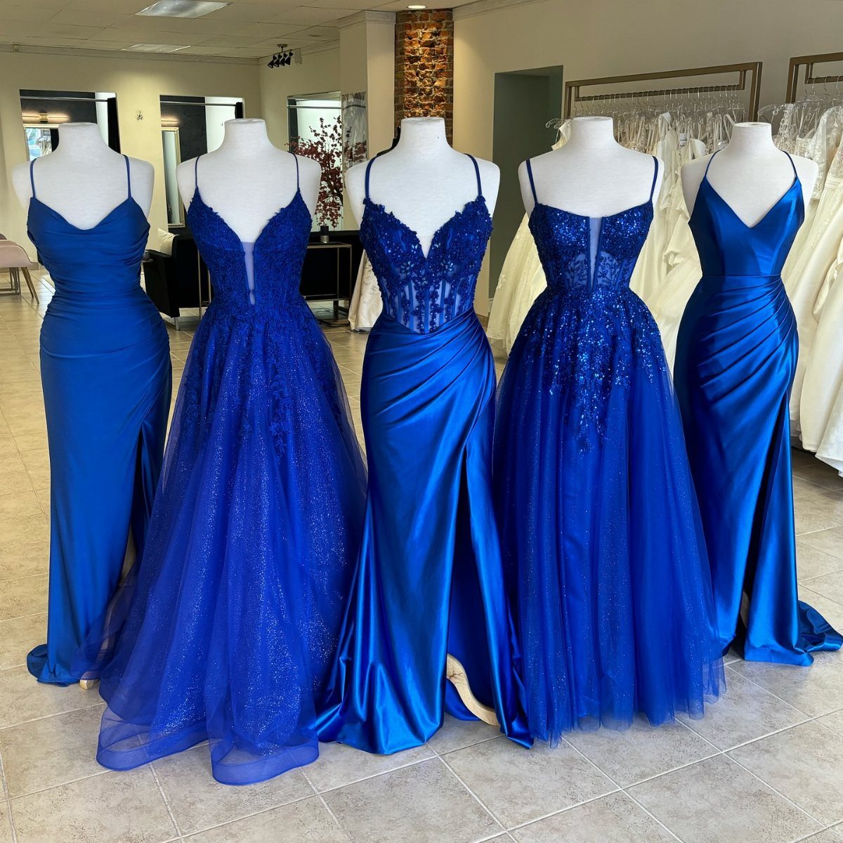 Five royal blue floor-length gowns displayed on white mannequins in a dress shop.