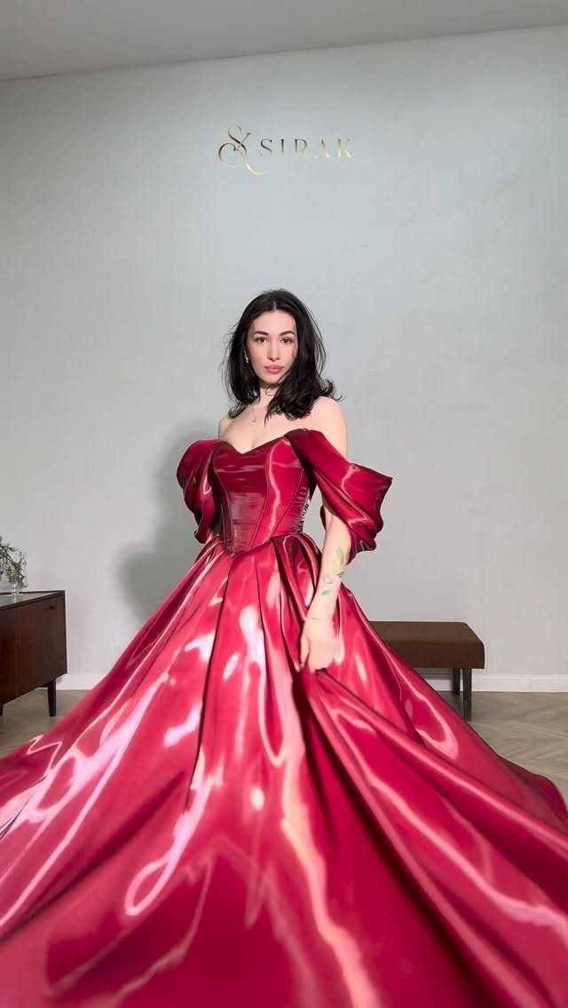 Woman wearing a voluminous, shiny red satin off-the-shoulder ball gown in a room.