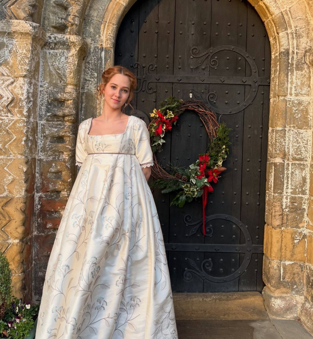 Woman in a cream floral satin empire-waist gown standing by a decorated door.
