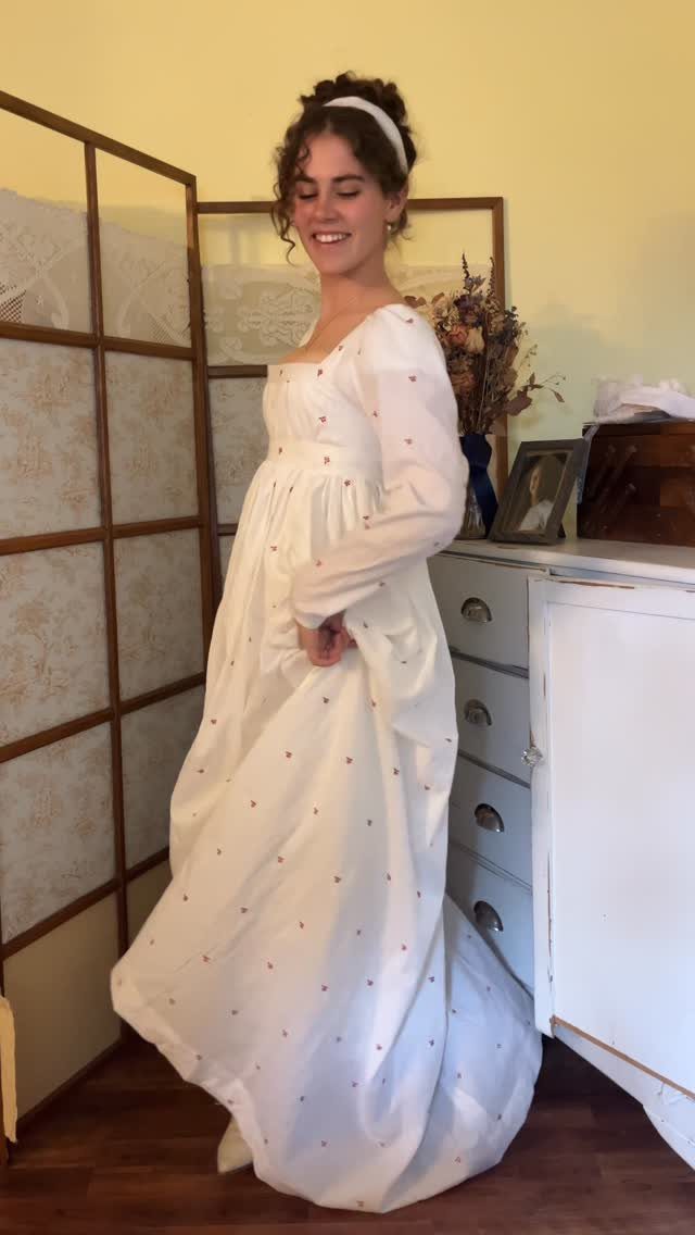 Woman in a white floral empire waist regency gown standing by a dresser.