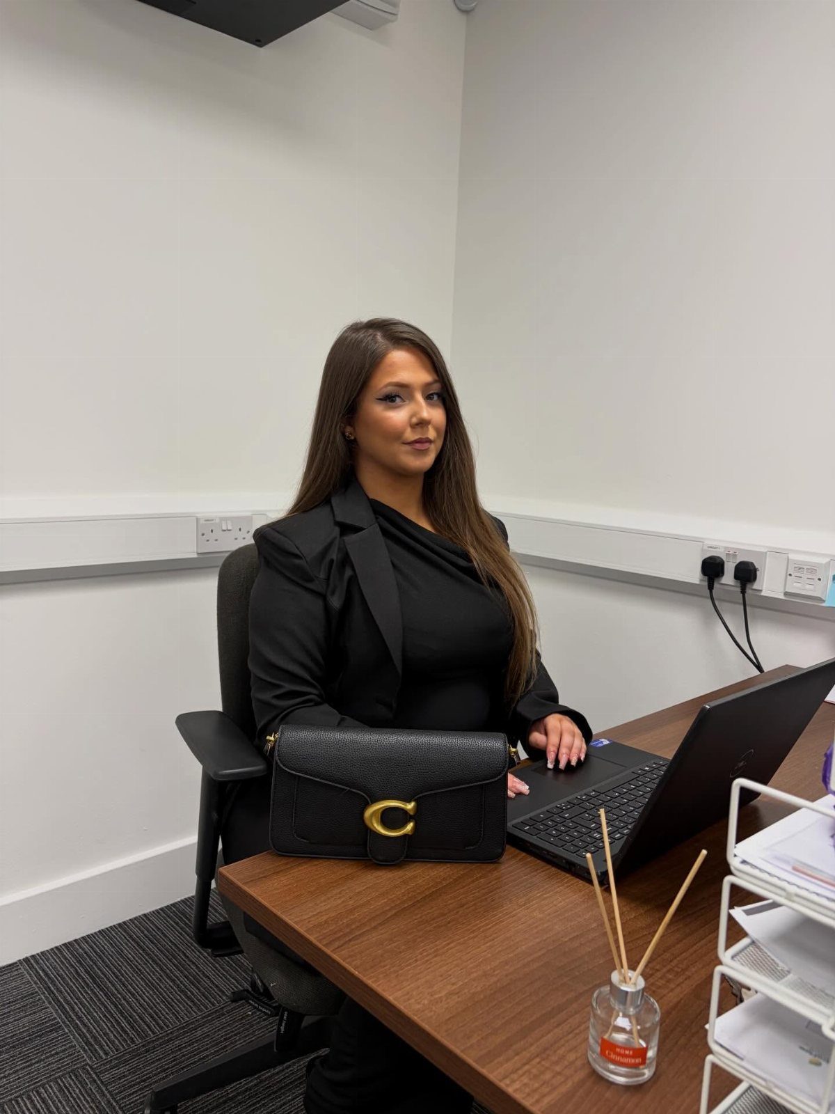 Woman in a black blazer and dress sitting at a wooden office desk