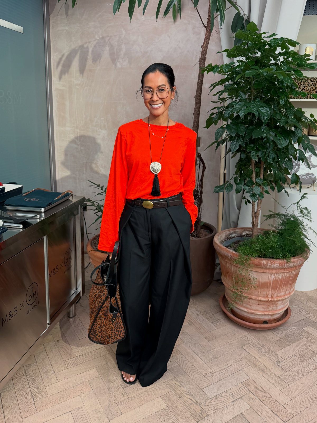 Woman wearing a bright orange blouse and black wide-leg tailored trousers indoors.