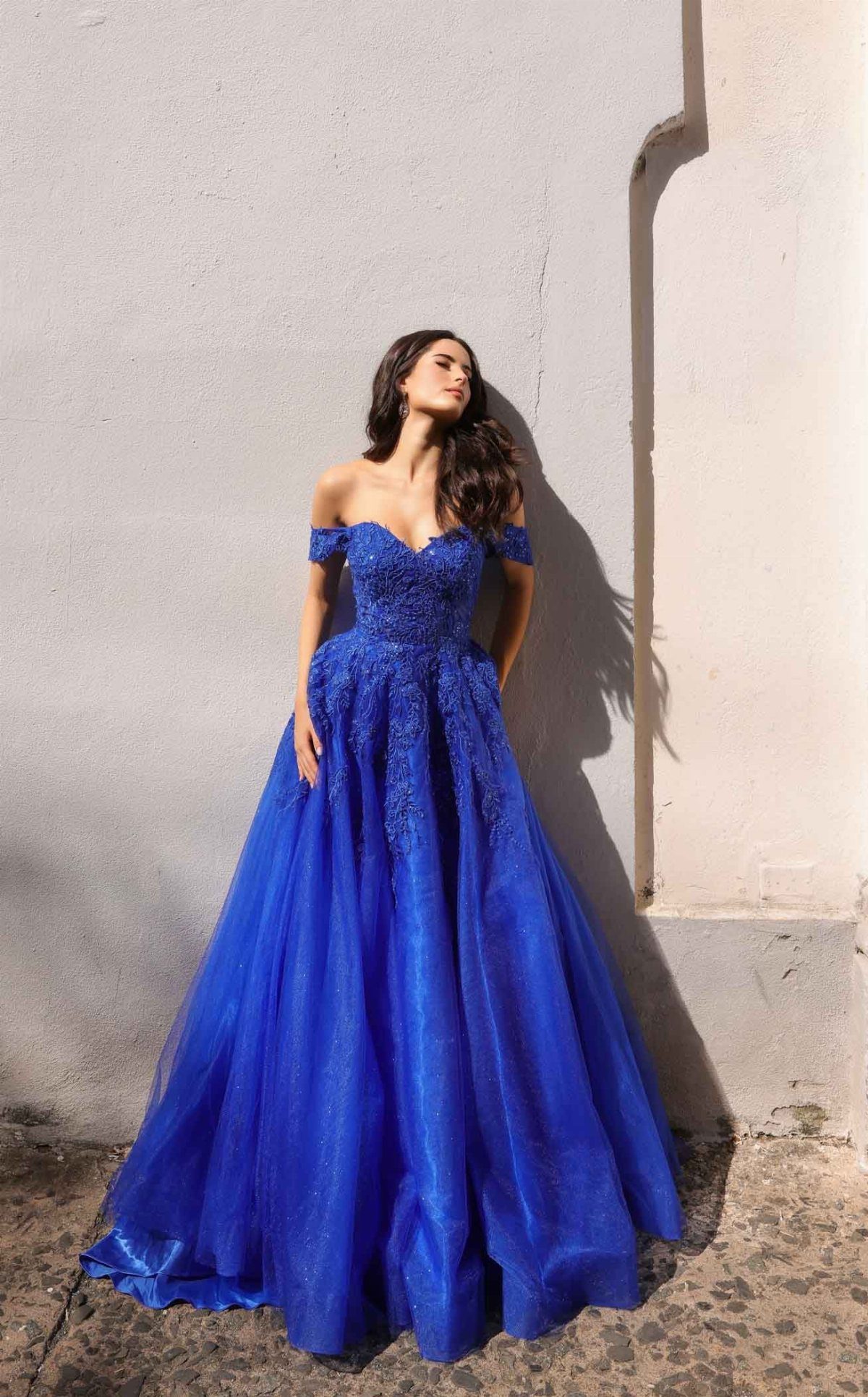Woman in royal blue off-the-shoulder tulle gown posing against a white wall