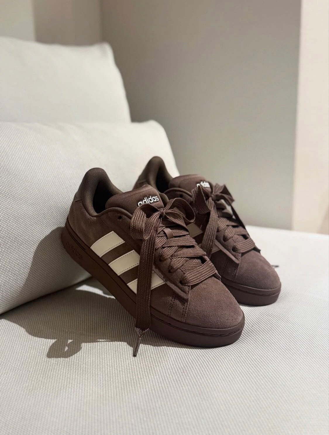 Brown suede Adidas sneakers with white stripes on a white fabric surface.
