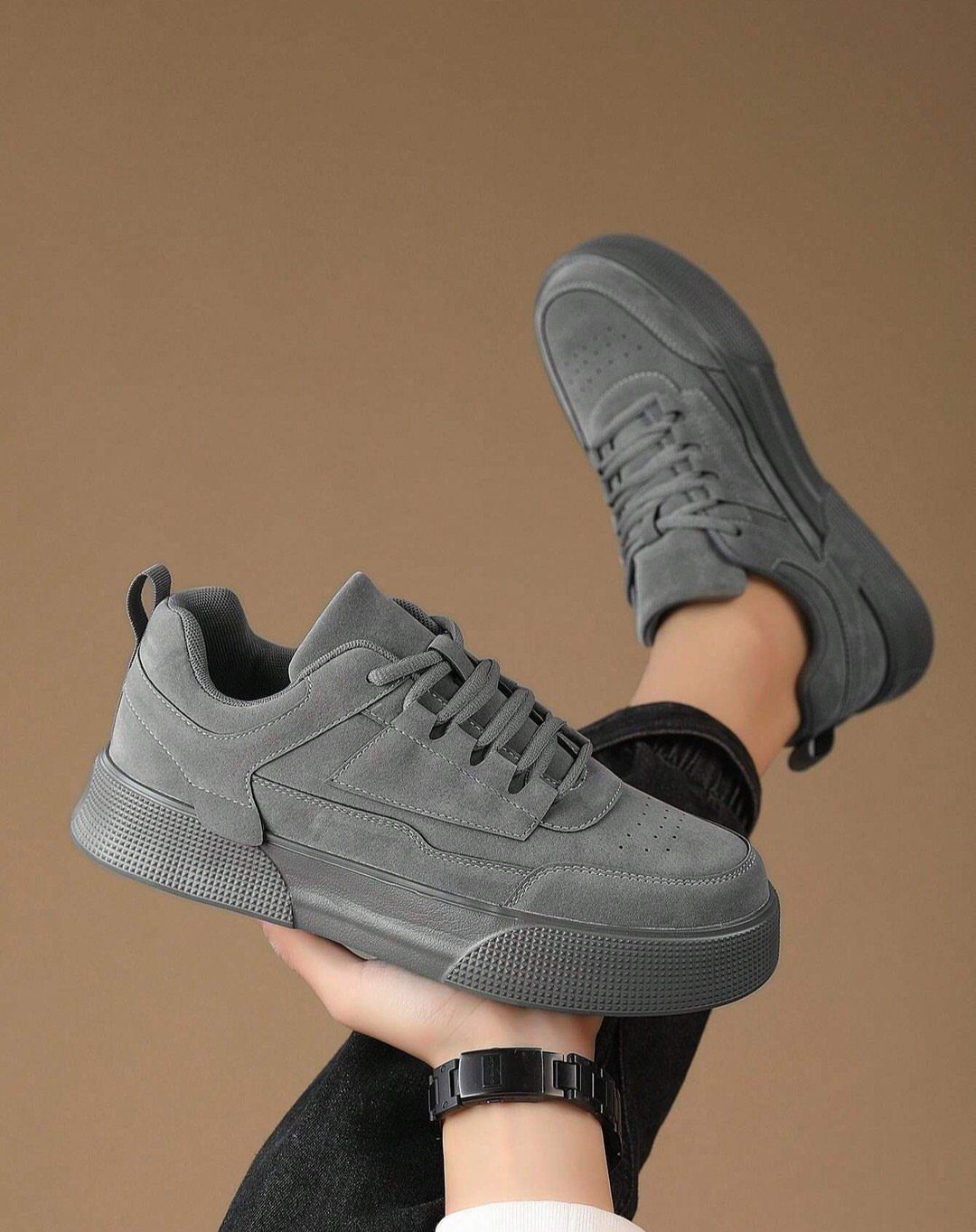 Gray suede sneakers held in hand and worn on foot against brown background