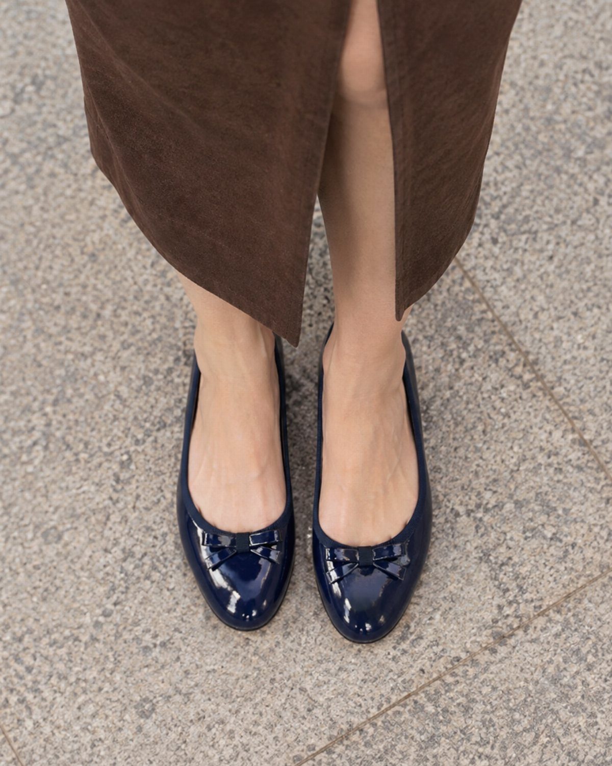 Navy blue patent leather ballet flats with bows worn with a brown skirt.