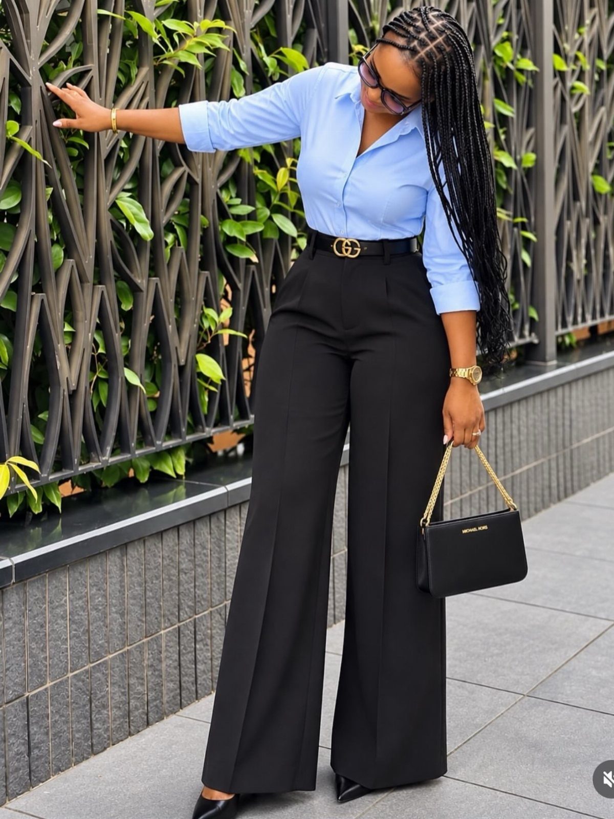 Woman in light blue shirt and black wide-leg trousers posing by a fence