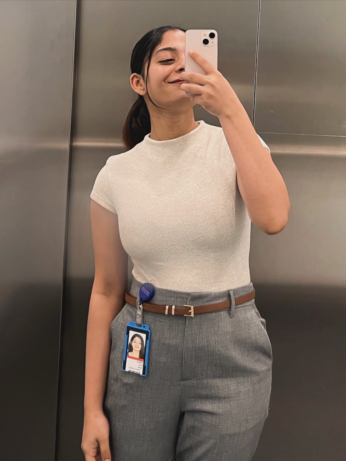 Woman in white t-shirt and grey trousers taking a mirror selfie in elevator