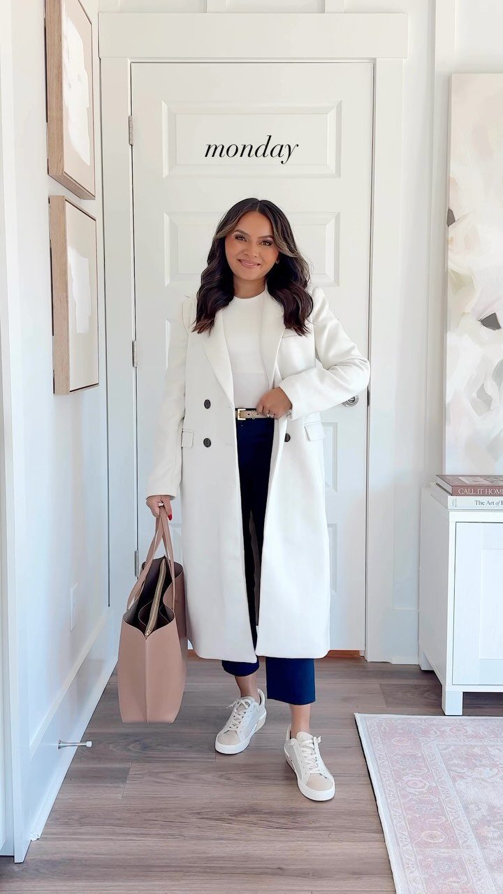 Woman in a long white coat and blue trousers standing in a hallway