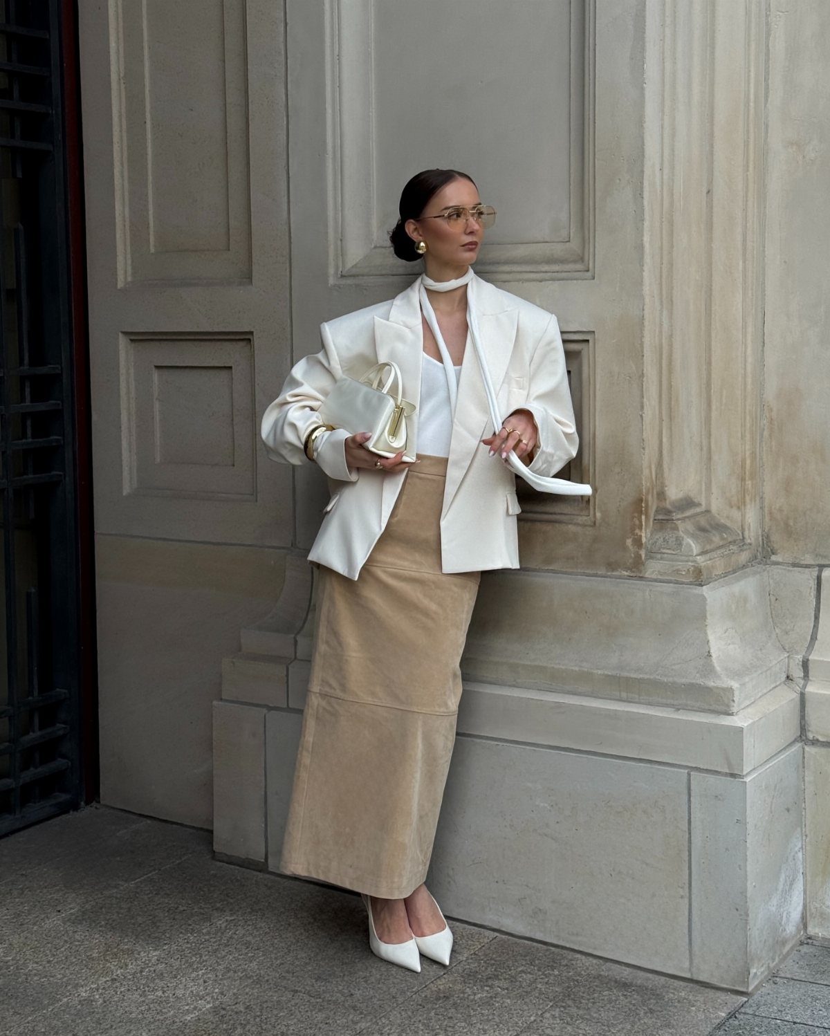 Woman in a white blazer and tan maxi skirt posing against a stone wall.