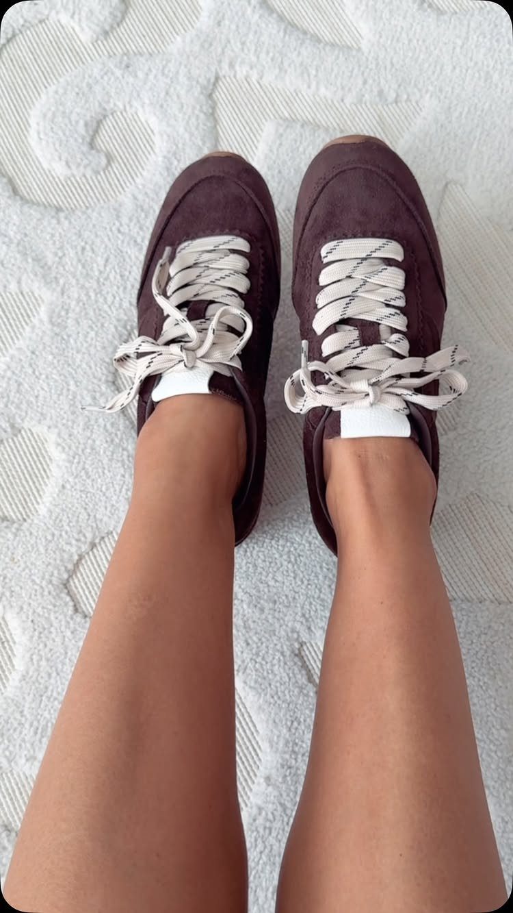 Burgundy suede sneakers with white laces worn by a person on white rug