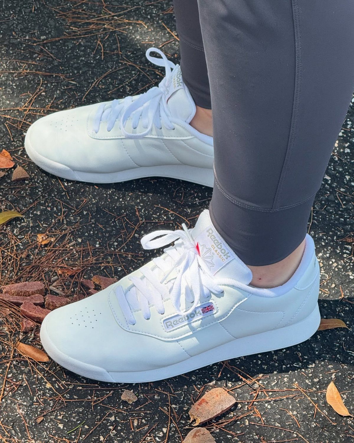 White Reebok sneakers worn with grey leggings on a leaf-strewn asphalt ground.