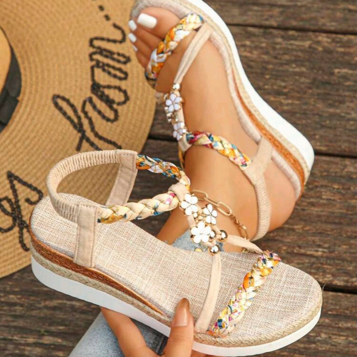 Beige sandals with braided colorful straps and white flower details on wooden decking.