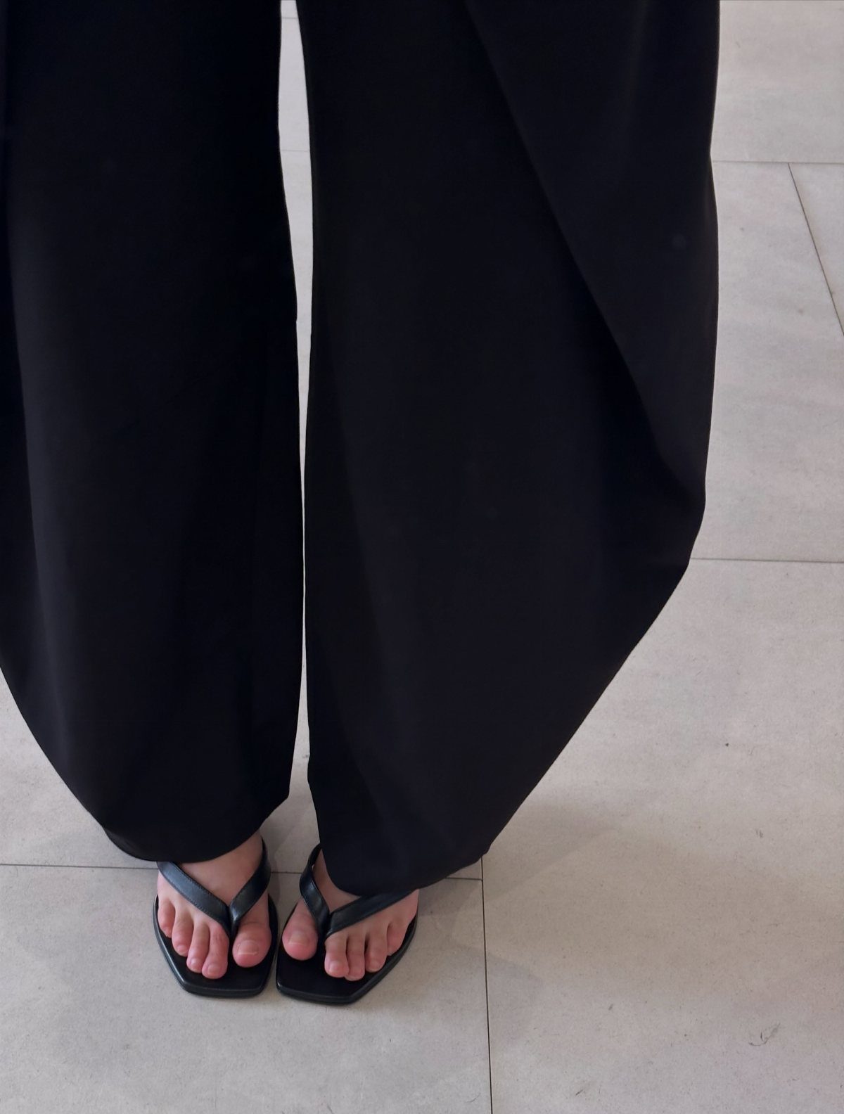 Black thong sandals with square toes worn with wide black trousers on tile.