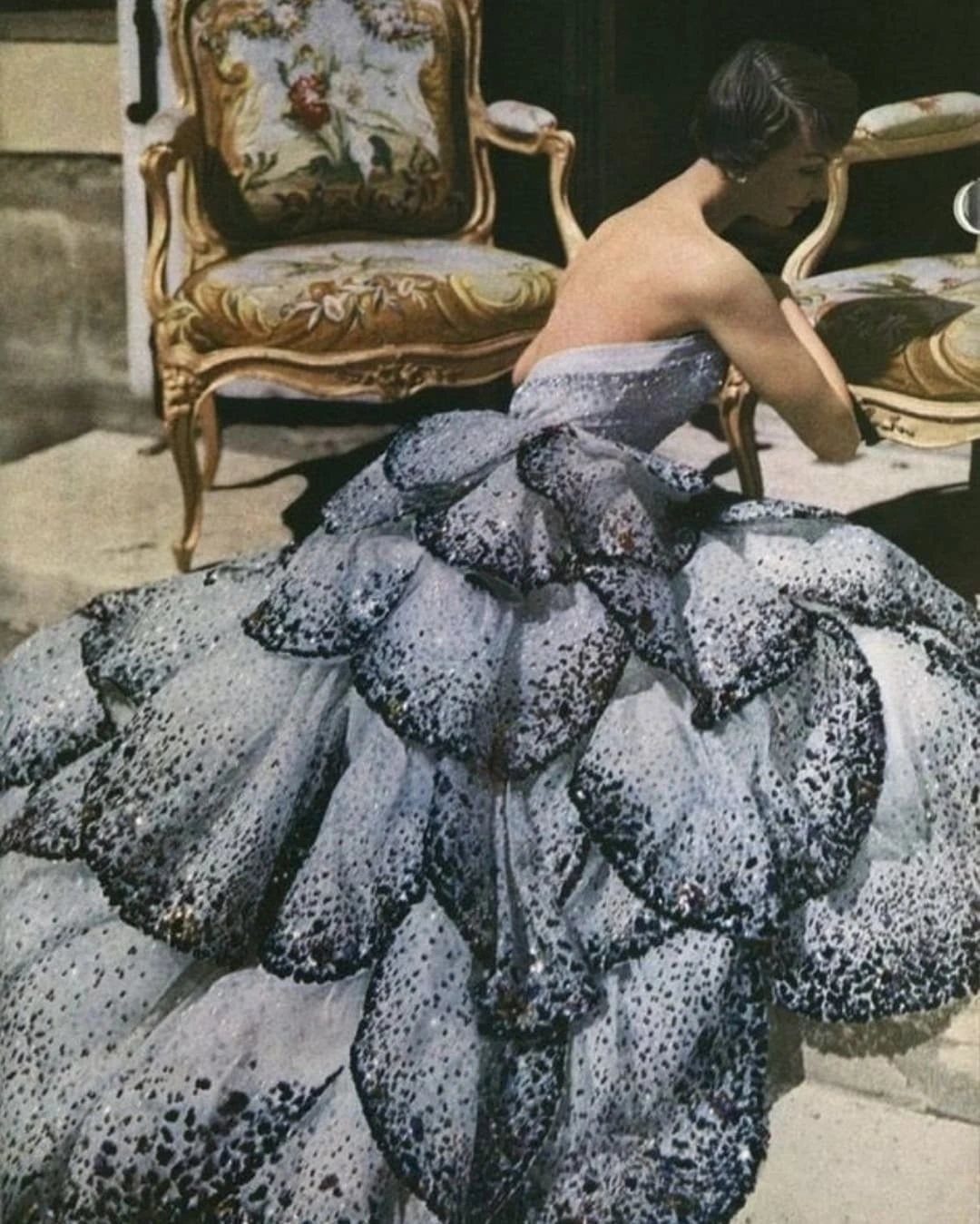 Woman in a voluminous grey petal-layered gown posing by ornate vintage chairs.